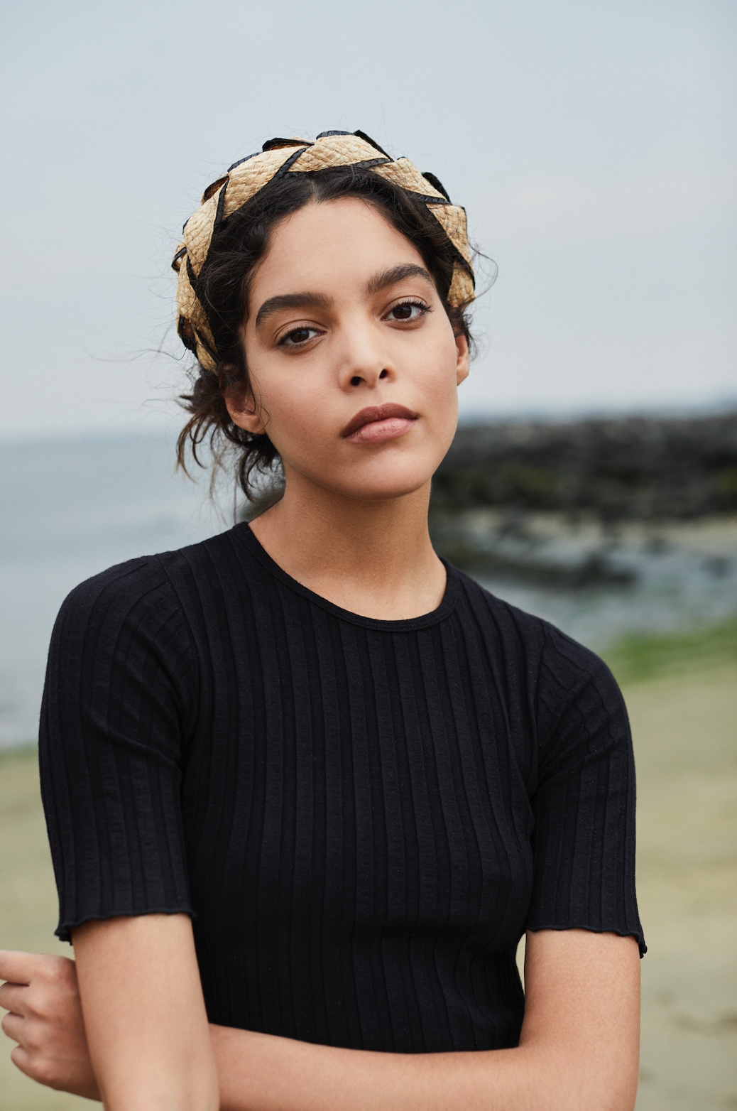 A person wearing a black shirt and the Gigi Burris Ava Band stands near a rocky beach, showcasing exquisite millinery craftsmanship.