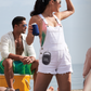 A woman wearing white overalls stands on a beach, gazing into the horizon while holding a drink. Nearby, a man sits as music fills the air from a JBL Clip 4 Waterproof Bluetooth Speaker by JBL, clipped to her shorts.