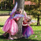 Two young girls play around a tree in a sunny park, wearing Great Pretenders Hot Pink Glitter Rainbow Wings as part of their fairy costumes.