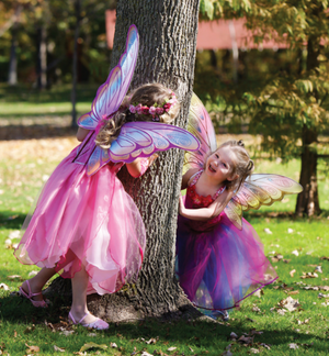 Two young girls dressed as fairies with Great Pretenders Hot Pink Glitter Rainbow Wings, playing hide and seek around a tree in a sunny park.