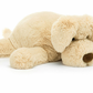 A large, light brown plush dog toy lies flat on its stomach with legs spread out and head resting on the ground, making the Jellycat Wanderlust by Jellycat the perfect chilled softie for any adventure.