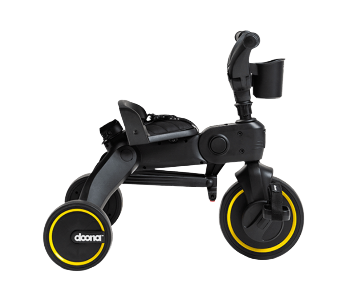 The DOONA Liki Trike - Midnight Edition is a black tricycle featuring a handlebar, seat, yellow-rimmed wheels, and a cup holder attachment. The brand name "Doona" is prominently displayed on the front wheel. This 5 in 1 compact folding trike serves as an excellent stroller alternative.