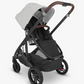 The UPPAbaby Cruz V2 Stroller by UPPAbaby comes in black and gray with a white canopy, brown handle, and an under-seat storage basket. It features four wheels, a reclining toddler seat, and an adjustable handlebar for added convenience.