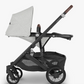 Side view of the UPPAbaby Cruz V2 Stroller in gray with a black frame, an adjustable handlebar, and brown accents. The stroller, by UPPAbaby, features four wheels, a storage basket underneath, and a comfortable toddler seat.