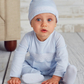 A baby in a light blue Kissy Kissy Footie with Hand Smocking and hat sits on a white wooden floor, looking up with a neutral expression. A beige piece of furniture is partially visible in the background.