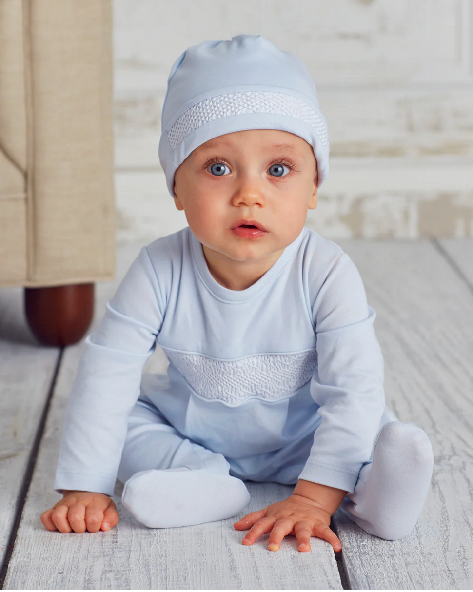 A baby in a light blue Kissy Kissy Footie with Hand Smocking and hat sits on a white wooden floor, looking up with a neutral expression. A beige piece of furniture is partially visible in the background.