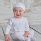 A baby wearing the Kissy Kissy Footie with Hand Smocking and a matching white pima cotton hat sits on a wooden floor, smiling and looking slightly to the side. The background features a painted, textured wall.