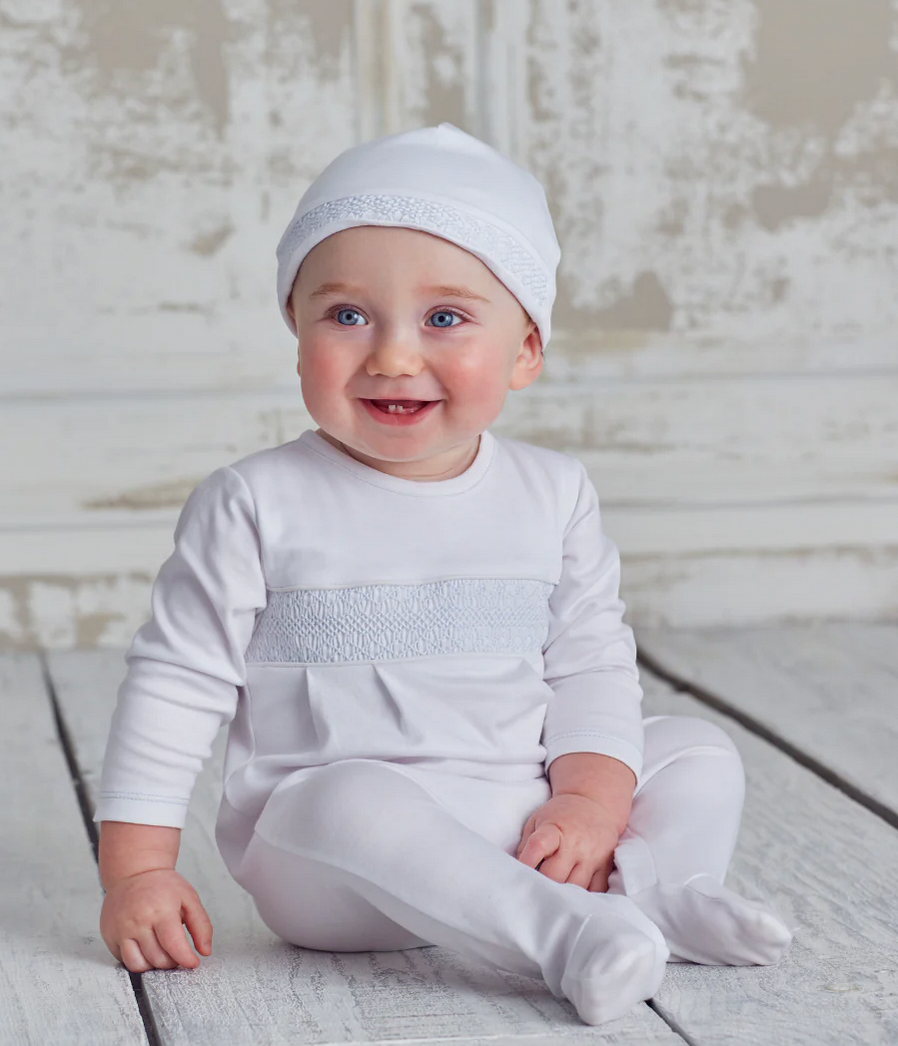 A baby wearing the Kissy Kissy Footie with Hand Smocking and a matching white pima cotton hat sits on a wooden floor, smiling and looking slightly to the side. The background features a painted, textured wall.