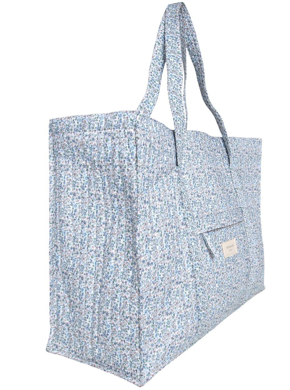 The Minnow Overnighter Tote by Minnow features a blue and white floral pattern, long handles, a front pocket, and a spacious interior crafted from quilted fabric.