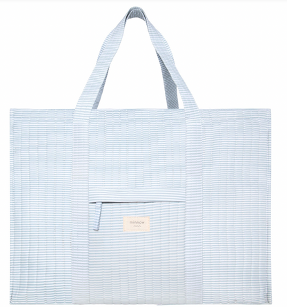 The Minnow Overnighter Tote by Minnow is a large blue striped quilted fabric bag featuring two handles, a front pocket with a small branded tag, and a spacious interior.