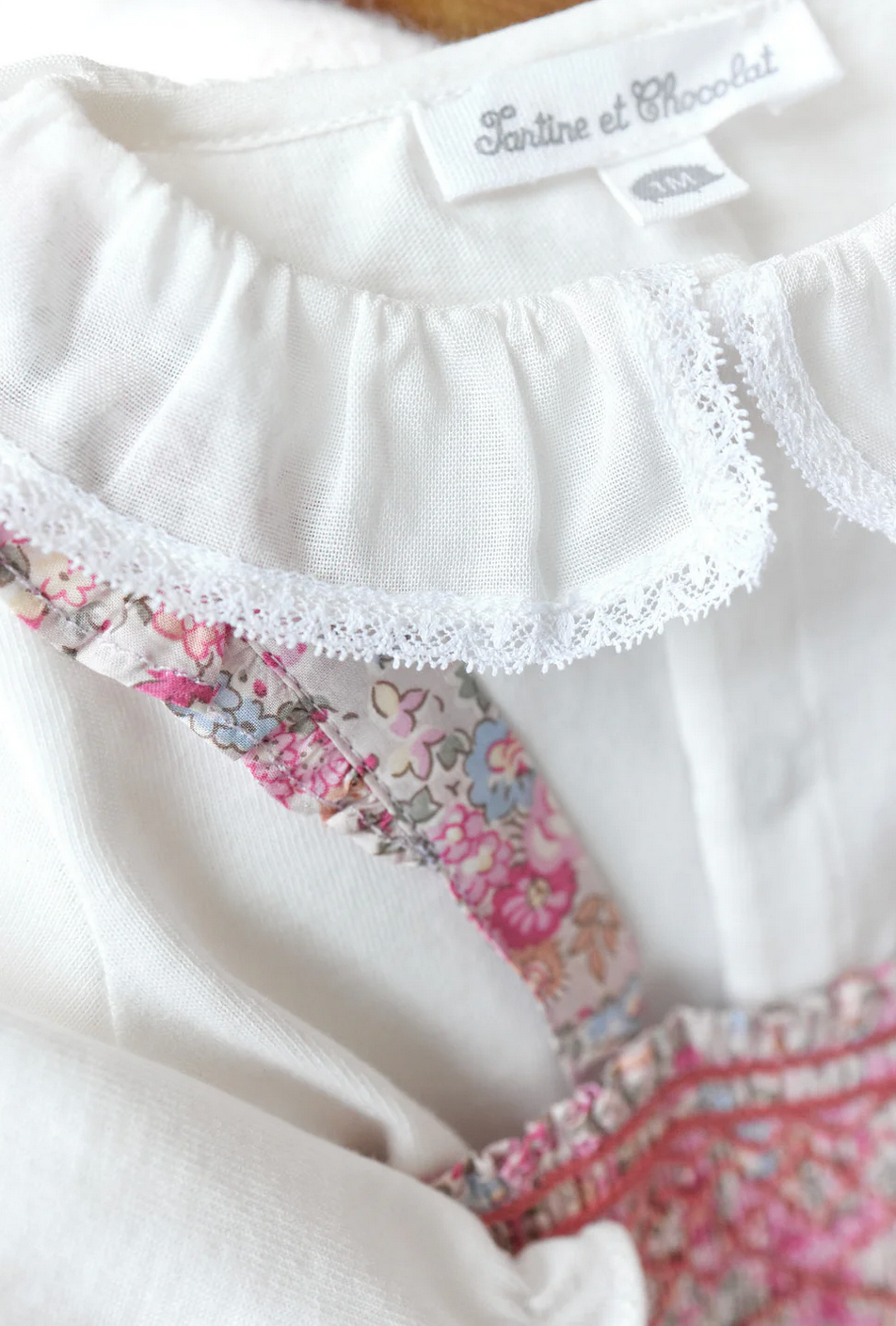 Close-up of the Tartine et Chocolat Ensemble Court Liberty Pink Overall Top Set, featuring a white garment with a ruffled collar. A pink floral-patterned fabric, likely Liberty cotton, peeks through the ruffles. The label reads "Tartine et Chocolat.