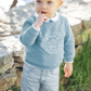 A blonde-haired baby boy stands outdoors in winter, wearing the Tartine et Choclat Camper Van Sweatshirt by Tartine et Chocolat and striped pants, with one finger in his mouth.