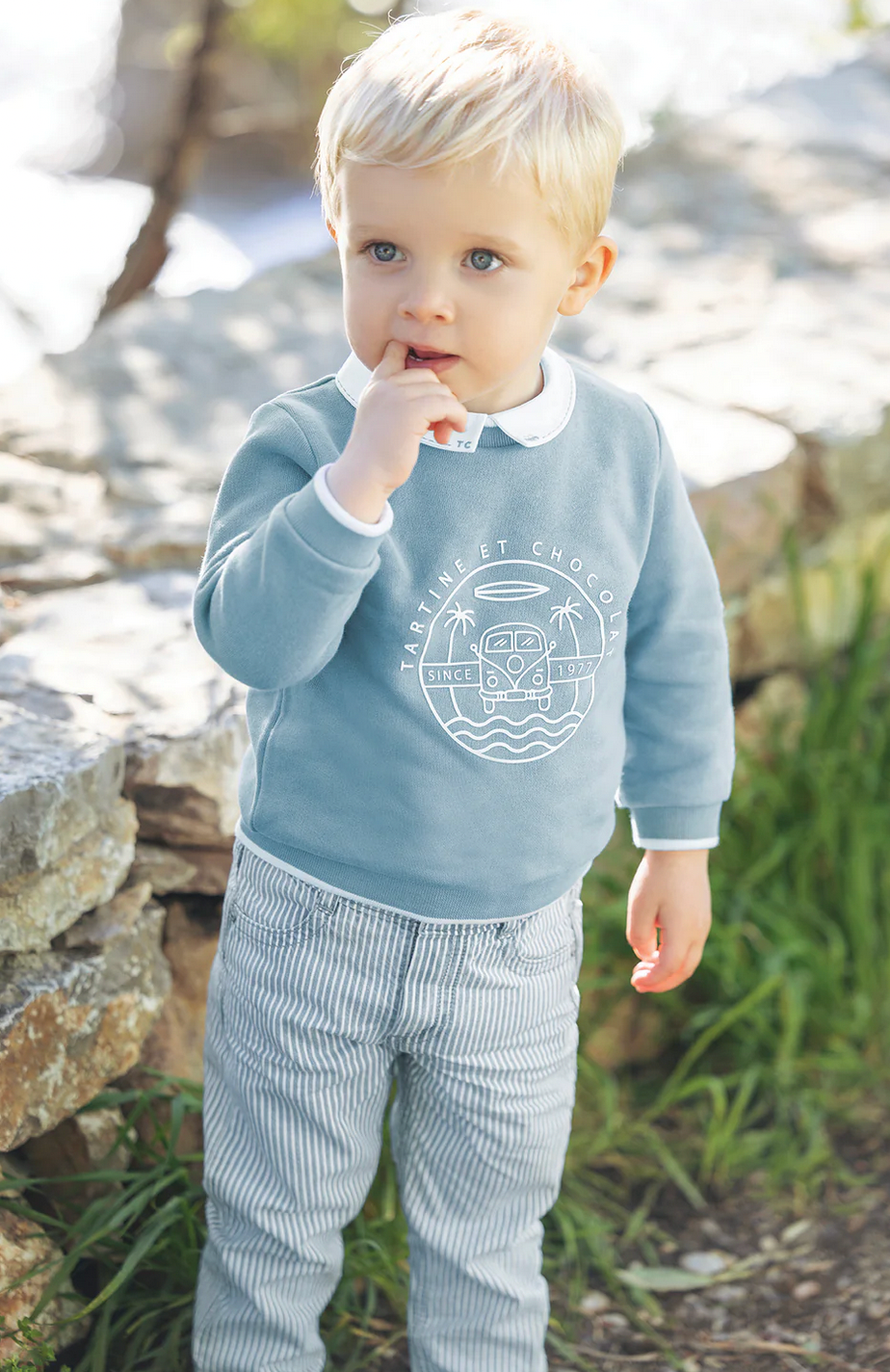 A blonde-haired baby boy stands outdoors in winter, wearing the Tartine et Choclat Camper Van Sweatshirt by Tartine et Chocolat and striped pants, with one finger in his mouth.