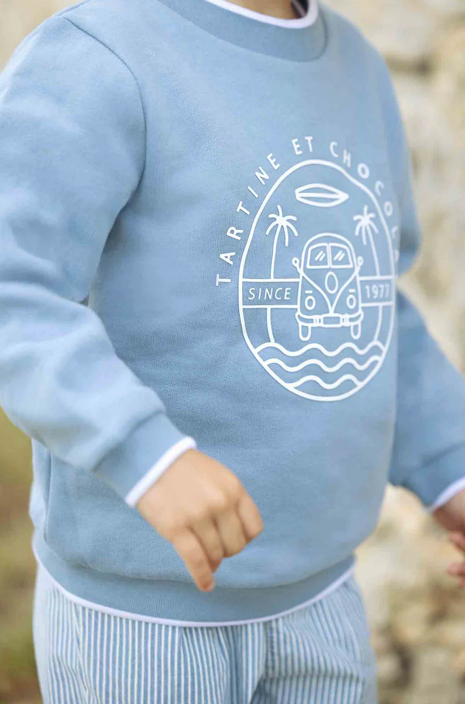 A baby boy is wearing the Tartine et Chocolat Camper Van Sweatshirt in blue, adorned with a white graphic of a van, palm trees, waves, and the text "Tartine et Chocolat Since 1977". He is also dressed in striped pants, making it ideal for winter.