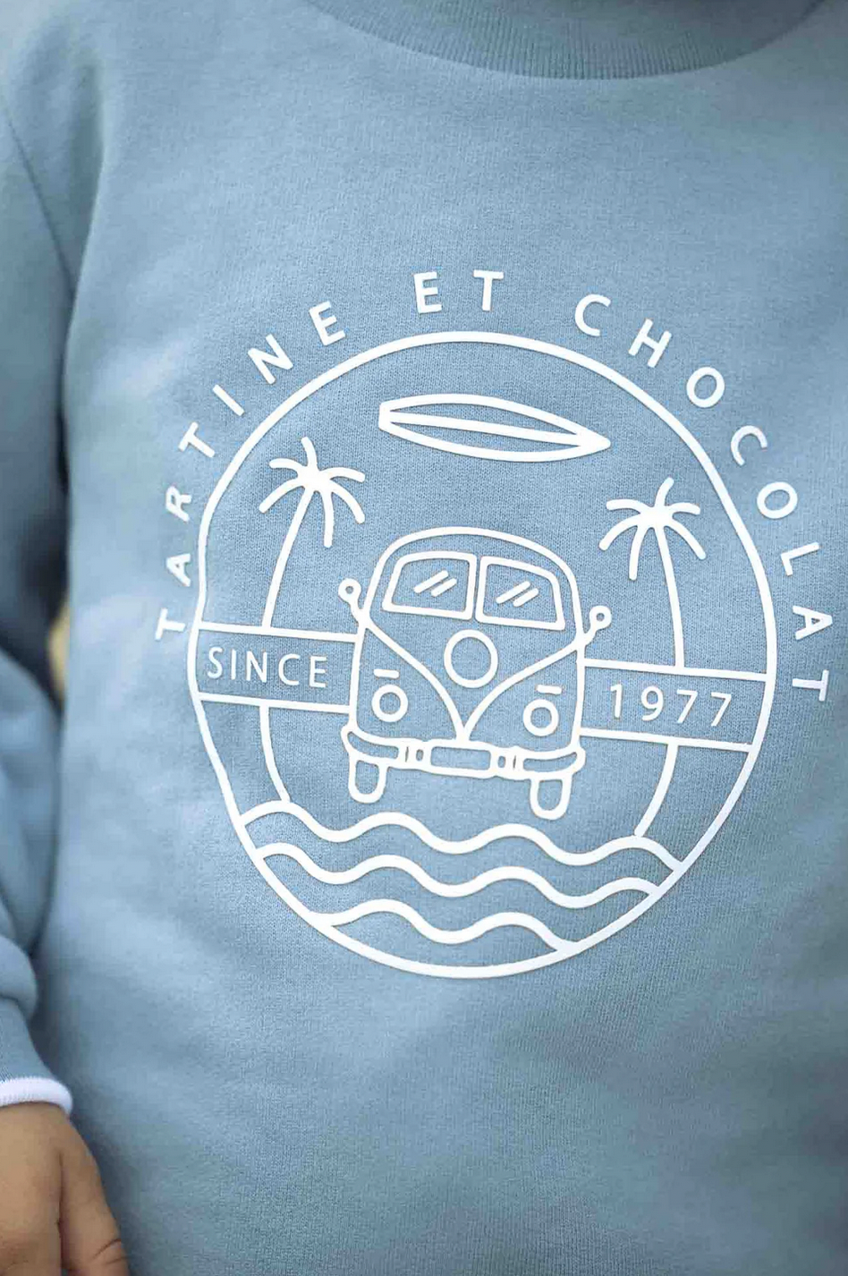 Close-up of the Tartine et Chocolat Camper Van Sweatshirt in blue, showcasing a logo with a van, palm trees, waves, and a surfboard, encircled by the text "Tartine et Chocolat" and "Since 1977." Perfect for your baby boy this winter.