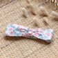 The Tartine et Chocolat Rasbery Printed Flowery Headband, featuring shades of pink, blue, and raspberry tones, is placed on a woven surface beside some dried wheat stalks.