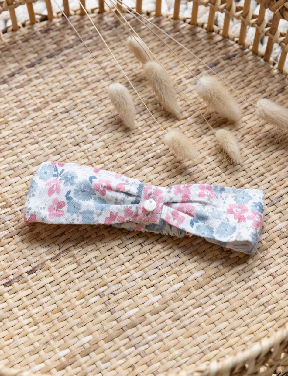 The Tartine et Chocolat Rasbery Printed Flowery Headband, featuring shades of pink, blue, and raspberry tones, is placed on a woven surface beside some dried wheat stalks.