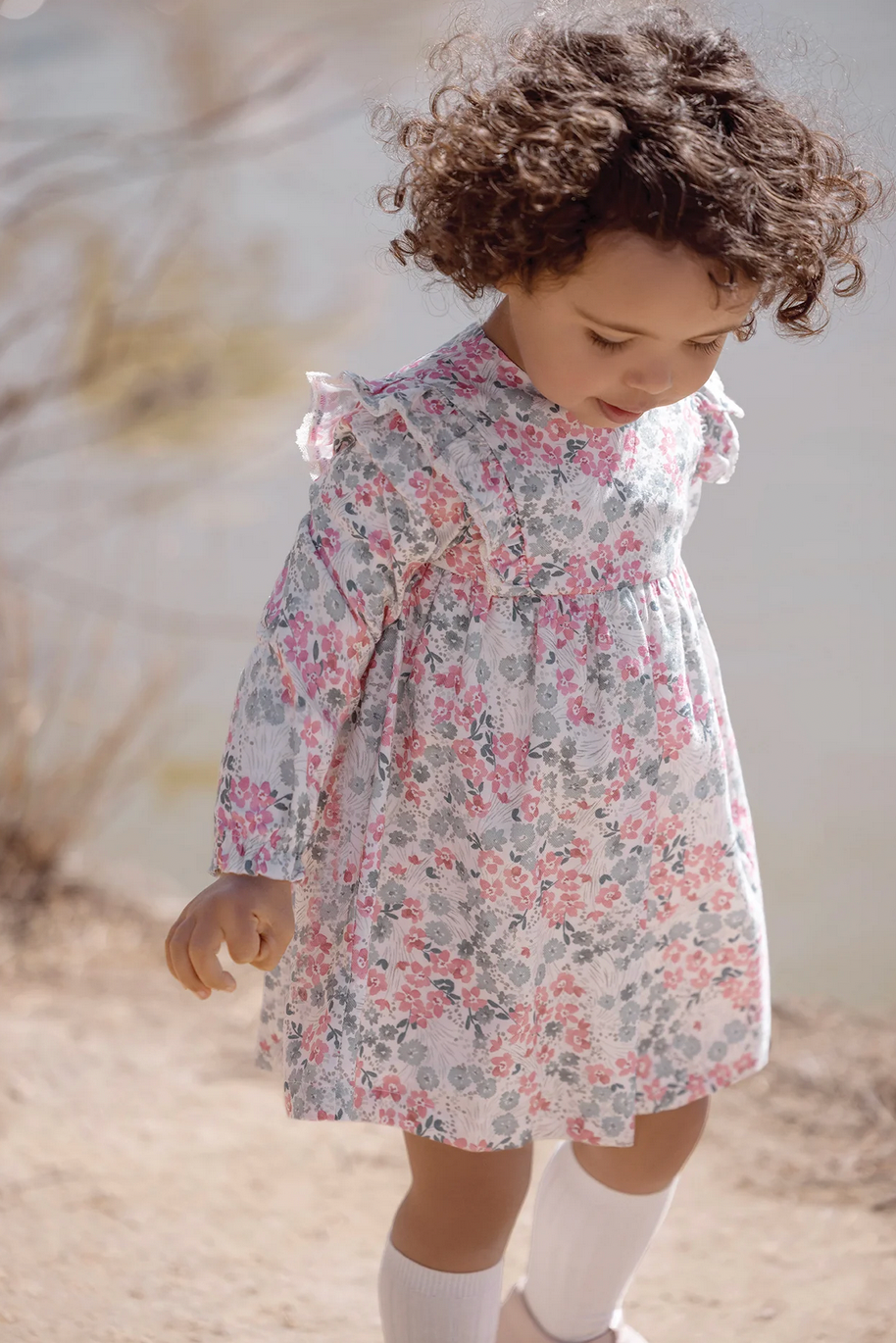 A young child with curly hair wears the Tartine et Chocolat Raspberry Printed Flowery Dress and looks down while walking outdoors.