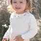 A young child with light hair is standing outdoors, wearing a Tartine et Chocolat Long Sleeve Onesie with an embroidered flower collar.