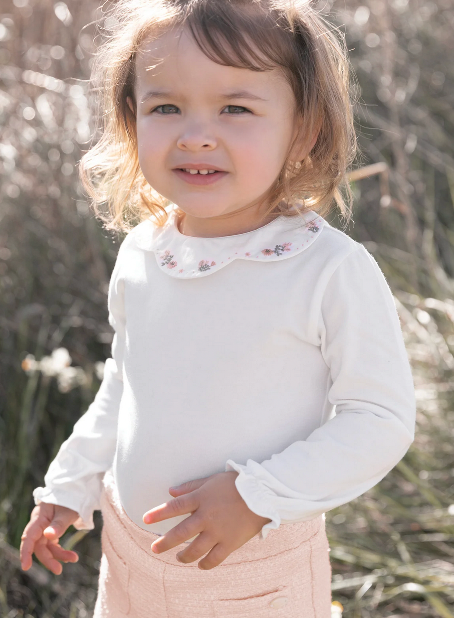 A young child with light hair is standing outdoors, wearing a Tartine et Chocolat Long Sleeve Onesie with an embroidered flower collar.