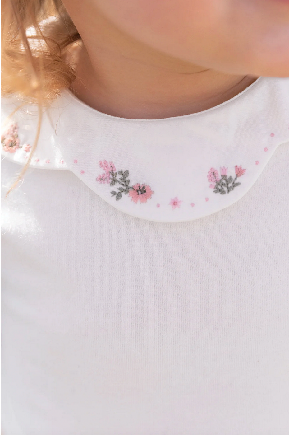 A close-up image of Tartine et Chocolat's Long Sleeve Onesie for children, showcasing a white shirt with a ruffled collar adorned with small pink and green embroidered flowers.