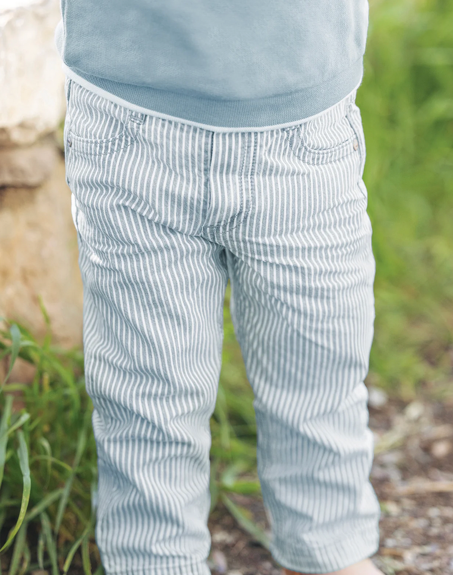 A toddler wearing the Tartine et Chocolat Sky Blue Striped Trousers stands outdoors near green grass and rocks, showcasing one of the many adorable outfits made from soft and comfortable fabric by Tartine et Chocolat.