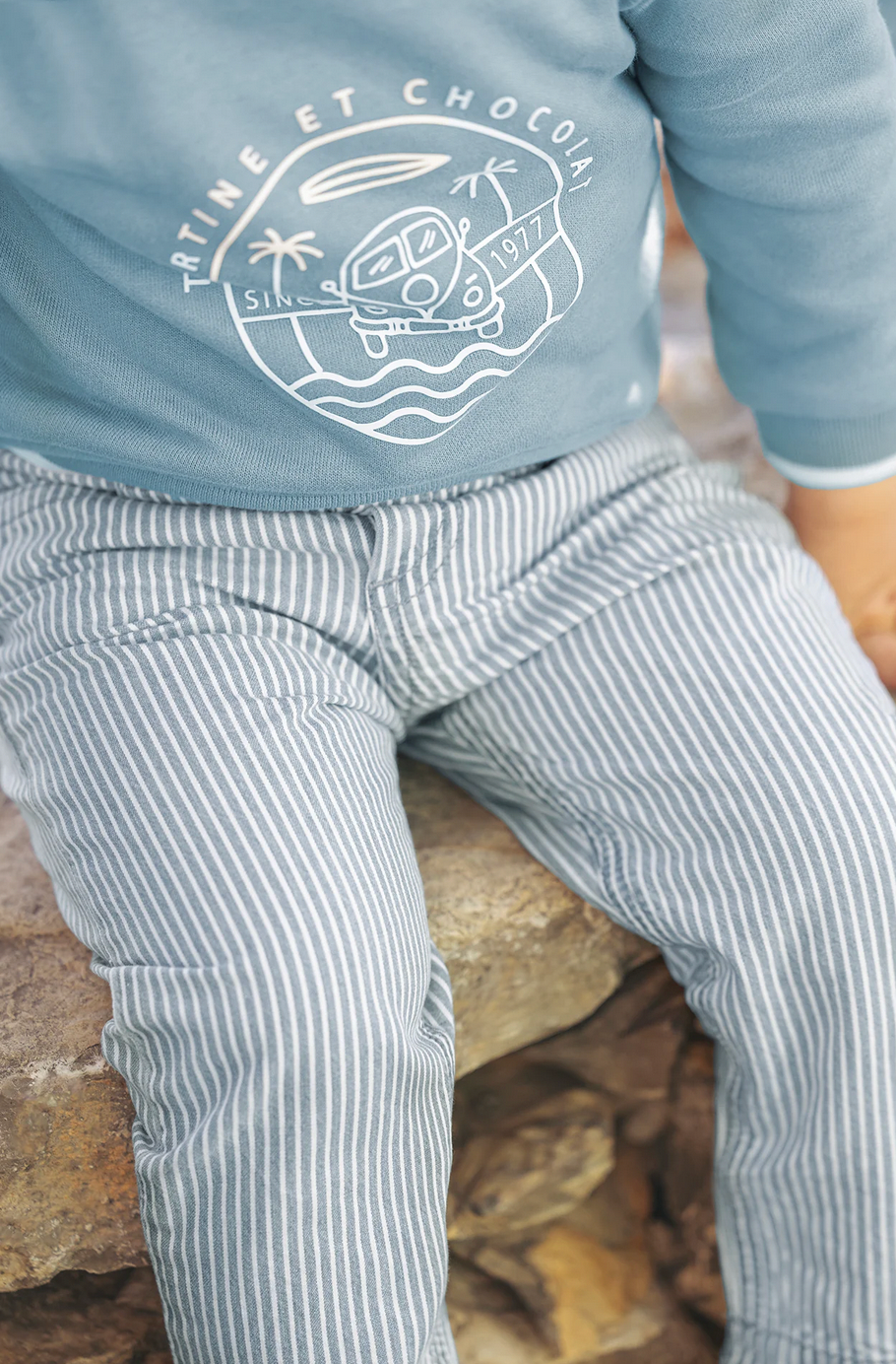 A child in an adorable outfit, featuring a light blue sweatshirt made of soft and comfortable fabric, paired with Tartine et Chocolat Sky Blue Striped Trousers for baby boys by Tartine et Chocolat, sits on a stone surface.