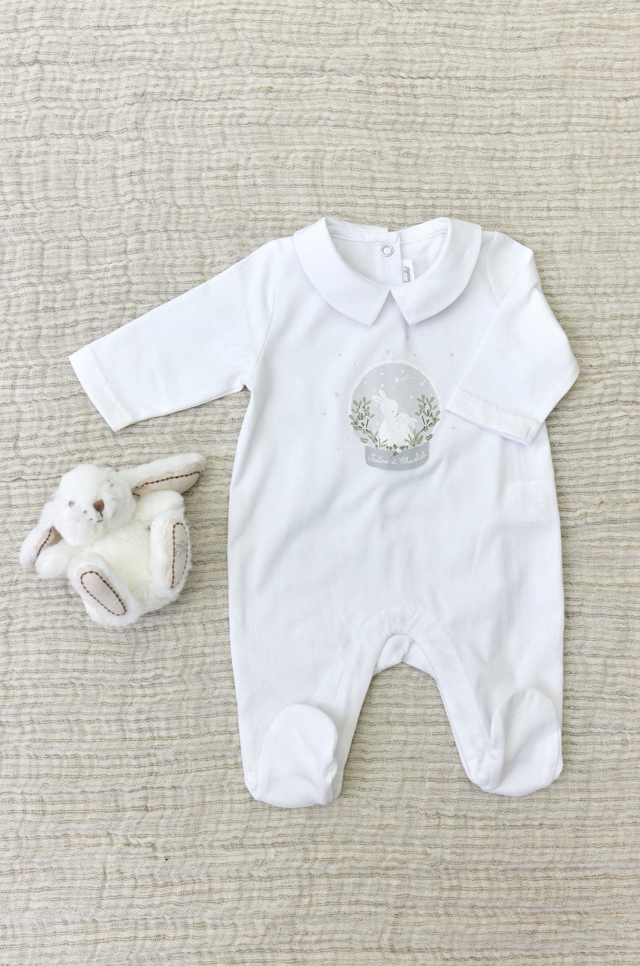 The Tartine et Chocolat Pyjama Snowball Bunny Onesie, made of 100% cotton with a snow globe design, laid on a beige textured background next to a small stuffed lamb toy. Perfect for newborn pajamas.