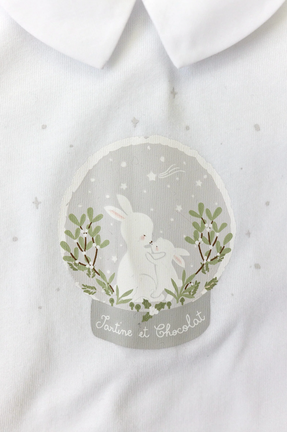 A close-up of the Tartine et Chocolat Pyjama Snowball Bunny Onesie shows a white 100% cotton fabric featuring an illustration of a larger and a smaller rabbit within a decorative circular frame, surrounded by stars and foliage, along with the text "Tartine et Chocolat" below. Perfect for newborn pajamas.