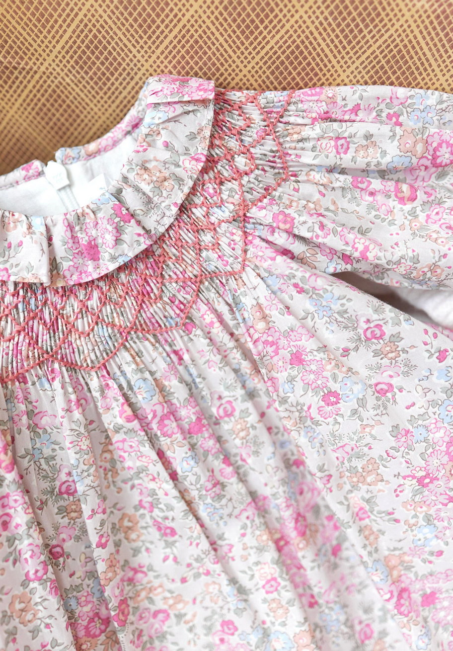 A close-up of the Tartine et Chocolat Fabric Liberty Pink Dress from Tartine et Chocolat, featuring intricate smocked detailing around the collar and chest area, and showcasing a beautiful Print Liberty floral pattern with pink and beige flowers on a light fabric.