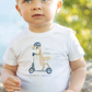 A young child stands outdoors wearing a comfortable Tartine et Chocolat Scooter Fox T-Shirt featuring a scooter-riding bear illustration, paired with striped pants, and looking slightly upwards.