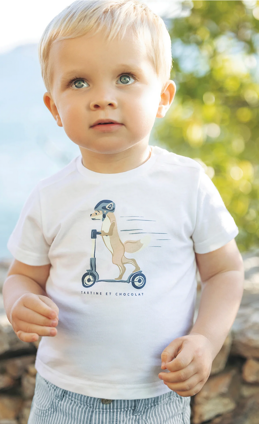 A young child stands outdoors wearing a comfortable Tartine et Chocolat Scooter Fox T-Shirt featuring a scooter-riding bear illustration, paired with striped pants, and looking slightly upwards.