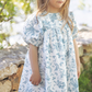 A young girl with long hair wears the Tartine et Chocolat Blue Printed Toile Dress, standing outdoors near a stone wall with greenery and sunlight in the background, perfect for special occasions.