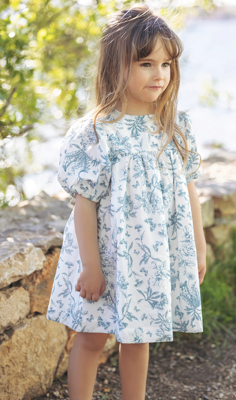 A young girl with long hair wears the Tartine et Chocolat Blue Printed Toile Dress, standing outdoors near a stone wall with greenery and sunlight in the background, perfect for special occasions.