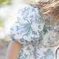 Close-up of a child's shoulder and arm wearing a white Tartine et Chocolat Blue Printed Toile Dress, with blue floral patterns reminiscent of toile de Jouy, and long brown hair partially visible. The background is blurred greenery.