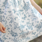 Close-up of a child's hand holding the hem of a Tartine et Chocolat Blue Printed Toile Dress by Tartine et Chocolat, featuring a delightful blue floral and butterfly toile de Jouy print, perfect for special occasions.