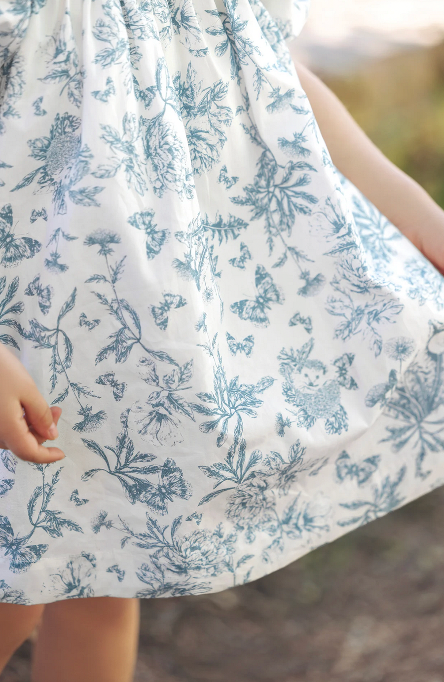 Close-up of a child's hand holding the hem of a Tartine et Chocolat Blue Printed Toile Dress by Tartine et Chocolat, featuring a delightful blue floral and butterfly toile de Jouy print, perfect for special occasions.