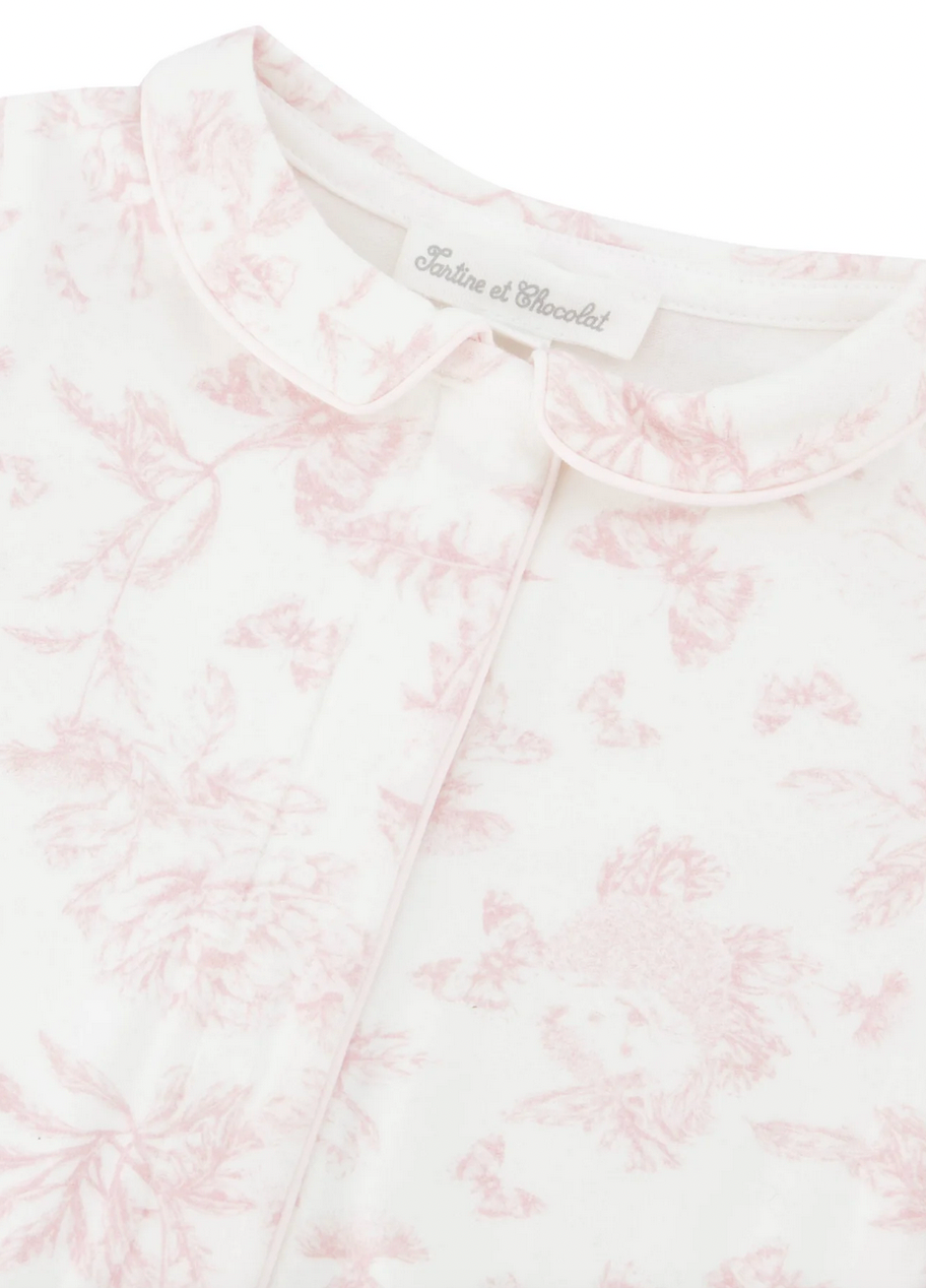 Close-up of the Tartine et Chocolat Pale Pink Toile Pyjama featuring a charming white and pink floral-patterned fabric with a Peter Pan collar. The soft and comfortable material boasts a label reading "Tartine et Chocolat," with pale pink hues enhancing its delightful design.