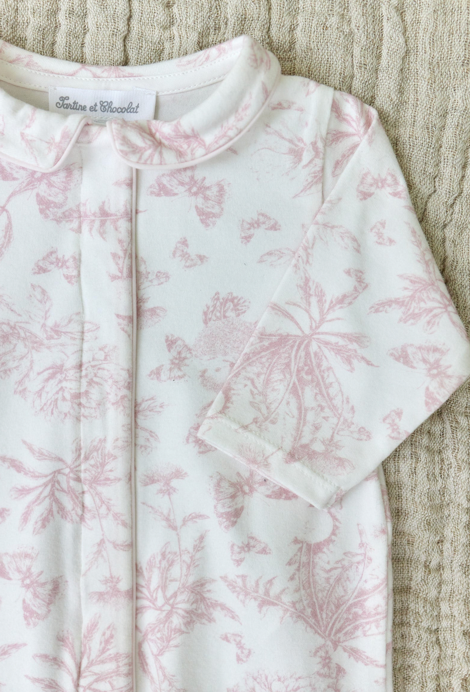 A close-up of the Tartine et Chocolat Pale Pink Toile Pyjama, featuring a white onesie adorned with pale pink floral patterns and a Peter Pan collar, lying on a beige textured surface—perfectly capturing the soft and comfortable fabric ideal for baby girl pajamas from the renowned brand, Tartine et Chocolat.