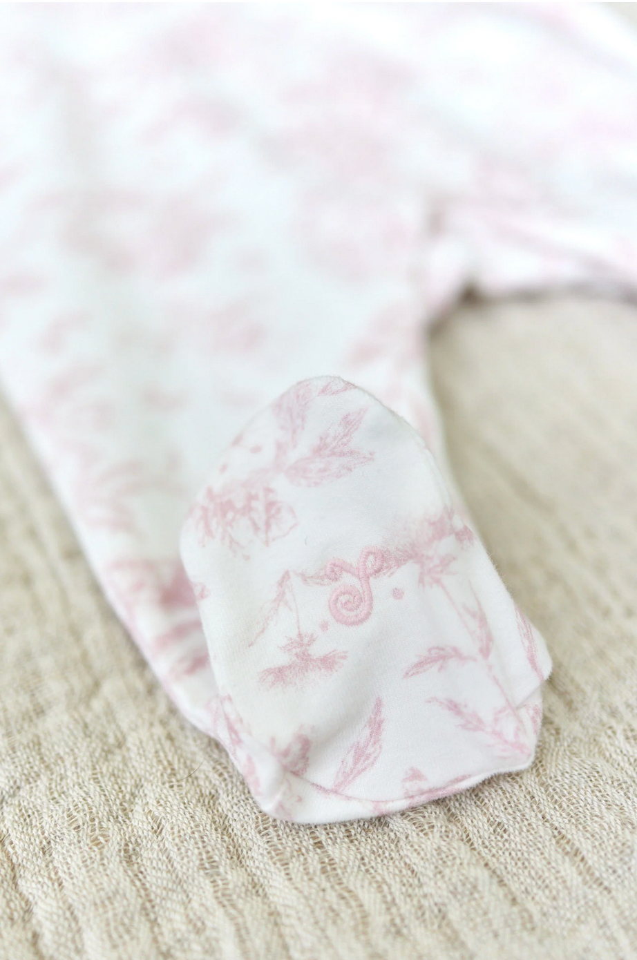 Close-up of a baby girl's foot covered by the Tartine et Chocolat Pale Pink Toile Pyjama, lying on a textured beige surface.