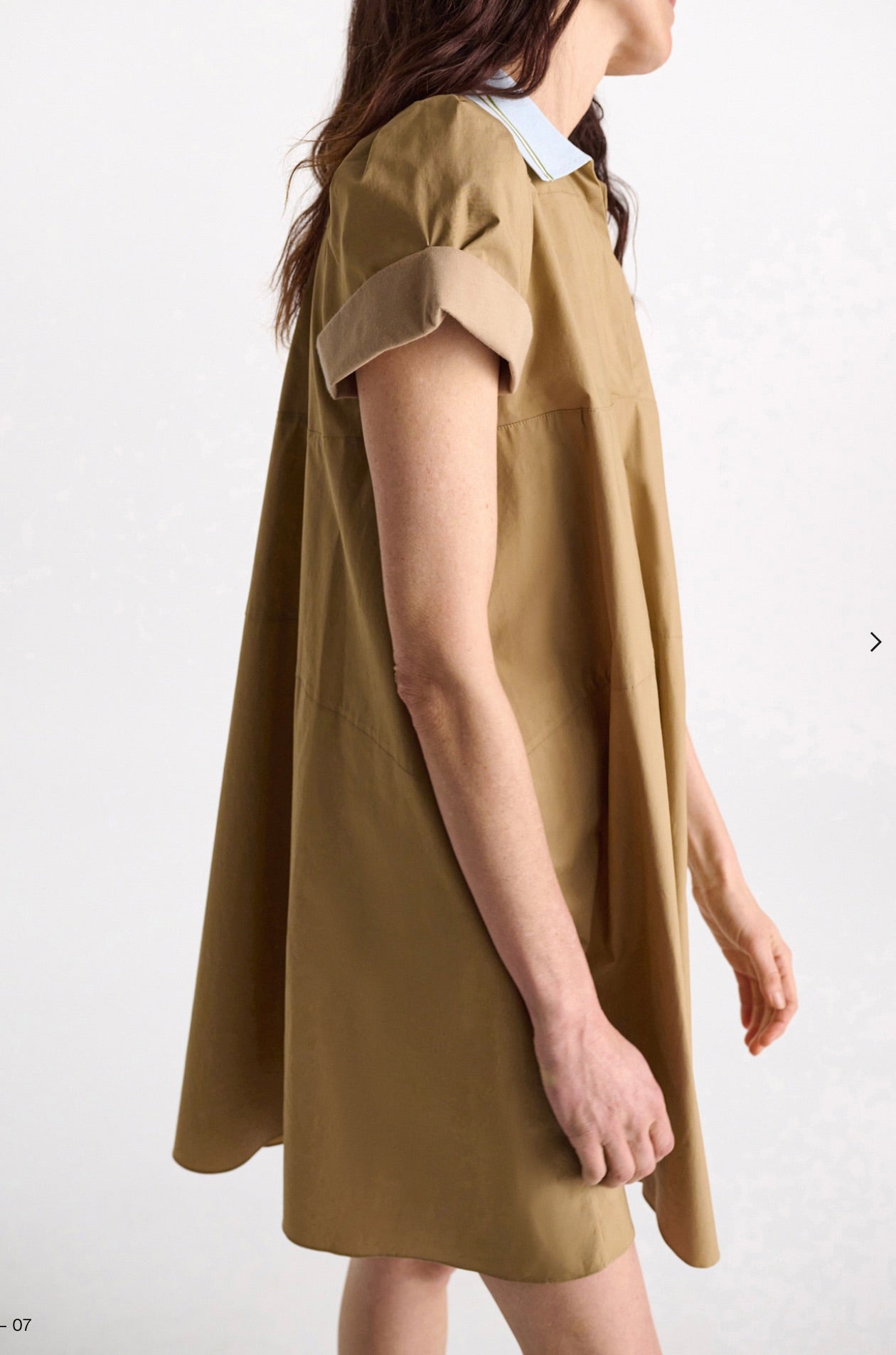 A woman models the Dorothee Schumacher Poplin Power Mini Shirt Dress, featuring a tan A-line cut, knee-length hem, short sleeves, and a light blue polo collar, as she stands facing slightly right against a plain background.