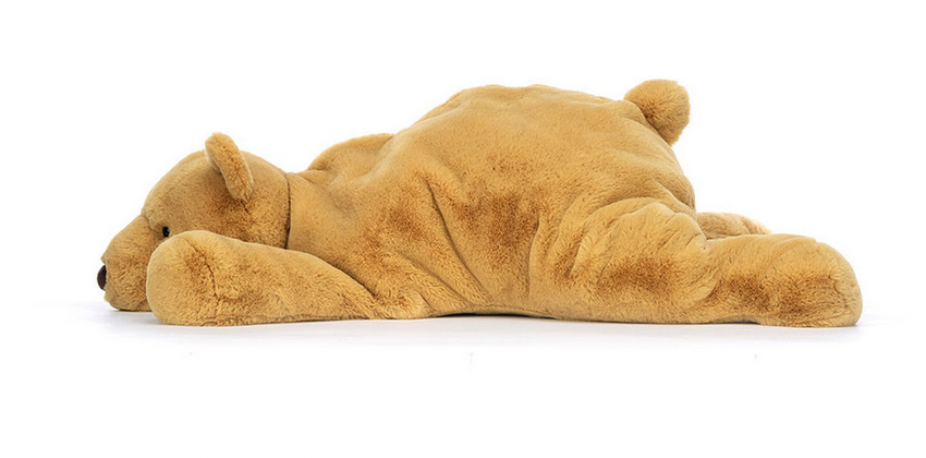 A large, light brown plush toy, specifically the Jellycat Harvey Bear from Jellycat, is lying on its stomach with its legs and arms outstretched.