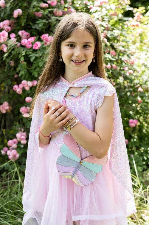 A young girl shines outdoors in the Great Pretenders Pink Sequins Twirl Cape, pairing it with a pastel butterfly purse—an enchanting addition to any children’s costume accessory collection.