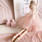 A MON AMI Belle Ballerina Doll by Mon Ami, dressed in a charming pink tulle dress and ballet slippers, sits gracefully on a bed. Behind it is a framed drawing of a perfume bottle, accompanied by two elegant pink decorative objects.