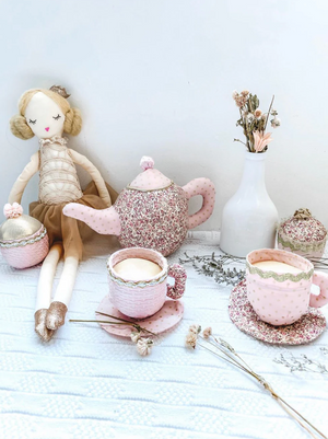 A Mon Ami Floral Stuffed Toy Tea Set sits with a cloth doll and a white vase of dried flowers, all arranged on a white textured surface.