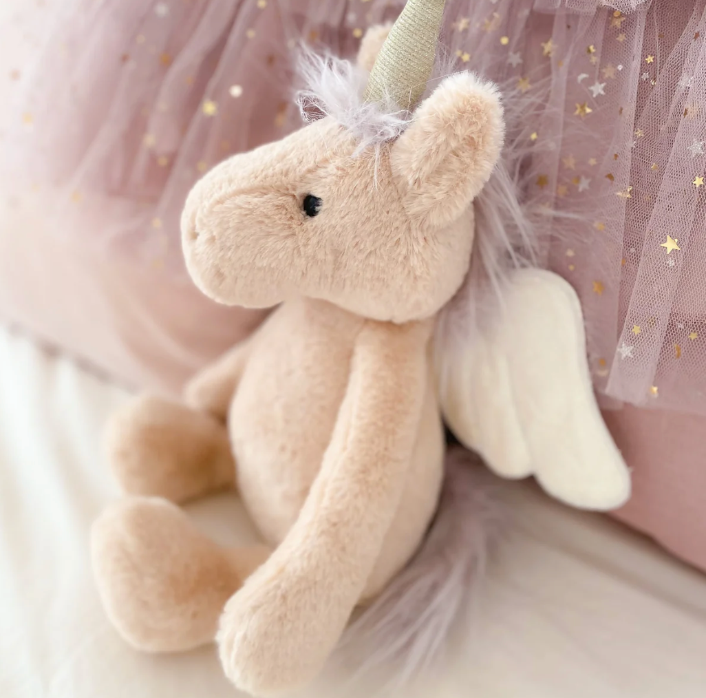 The MON AMI Luna Unicorn Fairy, a plush toy from Mon Ami featuring a horn and wings, sitting against pink tulle fabric adorned with gold stars, is the perfect gift for toddlers.