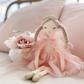 The Mon Ami Madeline Princess Doll, featuring braided hair and a pink tulle dress, sits on a bed surrounded by pale pink pillows and decorative pink and white flowers.