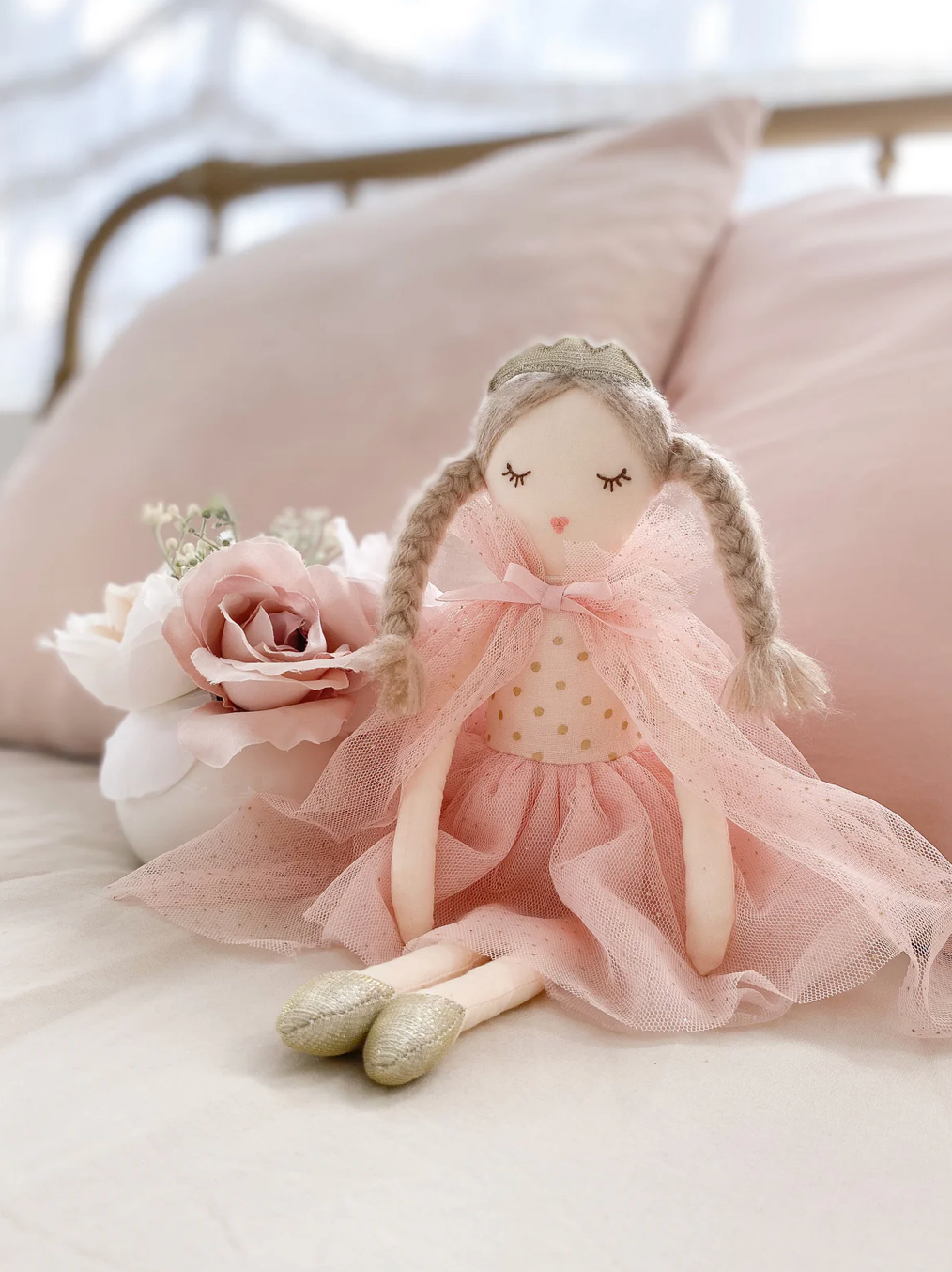 The Mon Ami Madeline Princess Doll, featuring braided hair and a pink tulle dress, sits on a bed surrounded by pale pink pillows and decorative pink and white flowers.