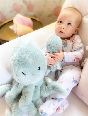 A baby in patterned pajamas sits on a white couch, holding the small Oda Octopus Plush Toy by Mon Ami—an adorable cuddle buddy—next to a larger matching plush, both perfect for baby shower gifts.