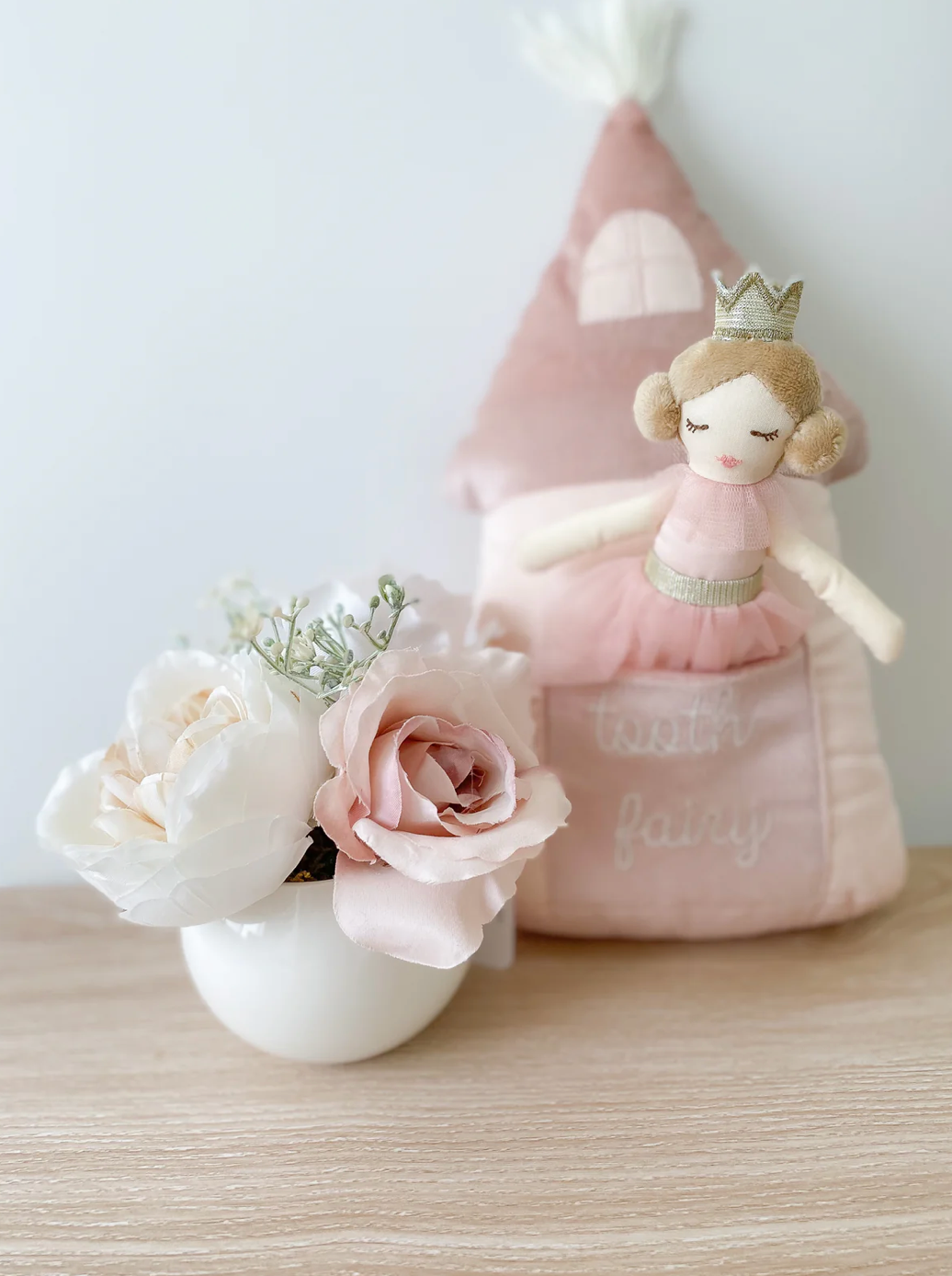 The Mon Ami Princess Castle Tooth Fairy Pillow Set is positioned next to a white vase filled with pink and white roses, creating a charming room decor setting on a wooden surface.